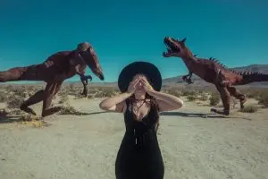 A woman covers her eyes amidst life-sized dinosaur sculptures in a desert setting.