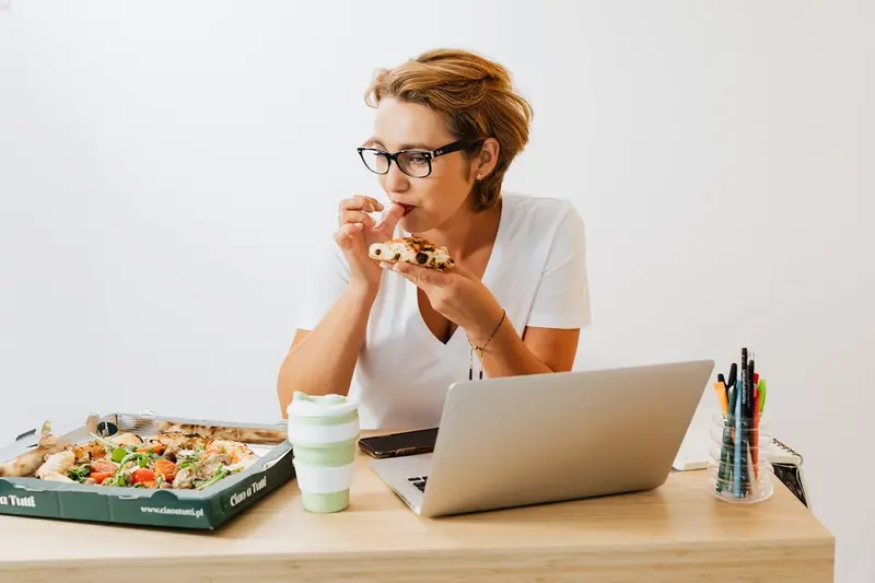 A woman by a laptop eating pizza